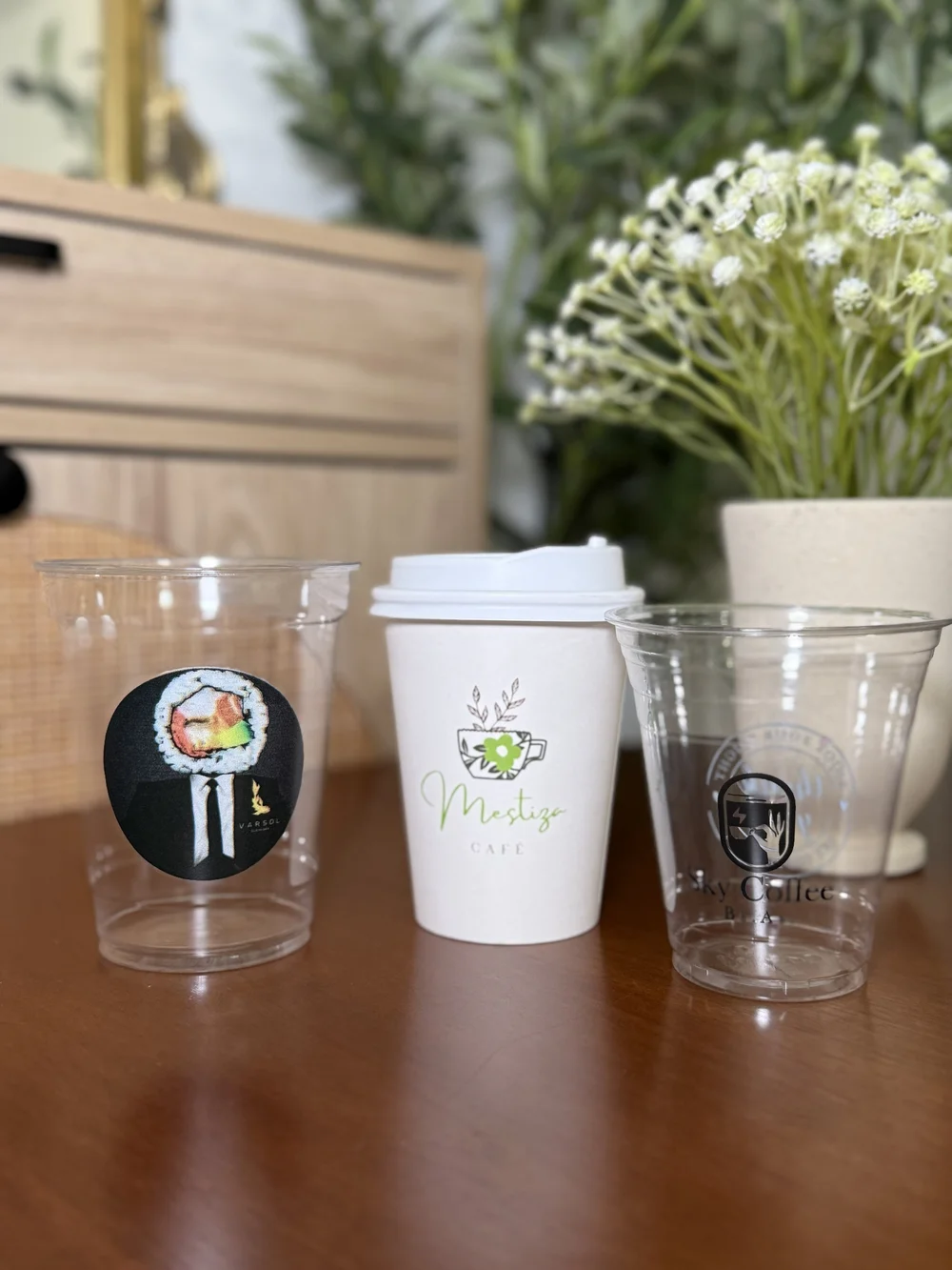 Three custom-printed cups on a wood table — a clear plastic cup with a sushi sticker, a white paper hot cup with lid printed with a "Mestiza Cafe" logo, and a clear plastic cold cup printed "Sky Coffee" — styled with flowers behind.