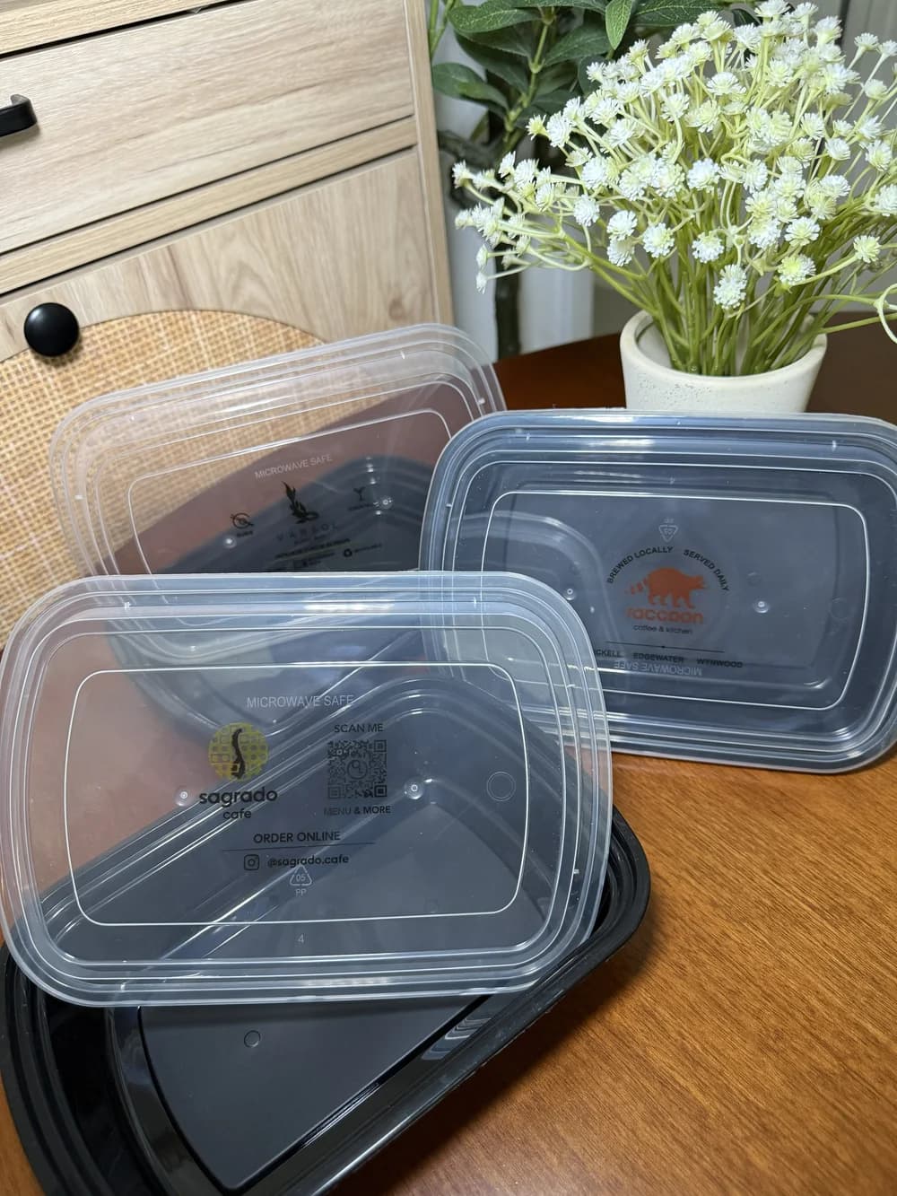 Real photograph of black microwave-safe takeout containers with clear plastic lids custom-printed with three different cafe logos, staged on a wooden table beside a flower pot.