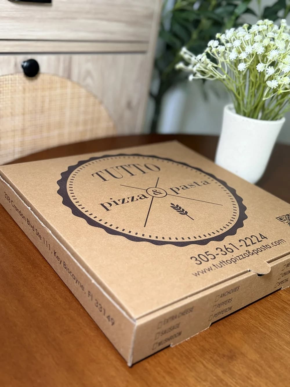 A kraft corrugated pizza box printed with a black circular Tutto Pizza & Pasta logo, photographed on a wooden restaurant table beside white flowers.